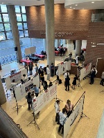 A downward view of 6 academic poster holders in a large open atrium with groups of people standing around the posters talking.