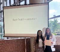 A picture of two women who are standing in front of a projector screen that reads Best Oral Presentation. One woman holds a certificate.